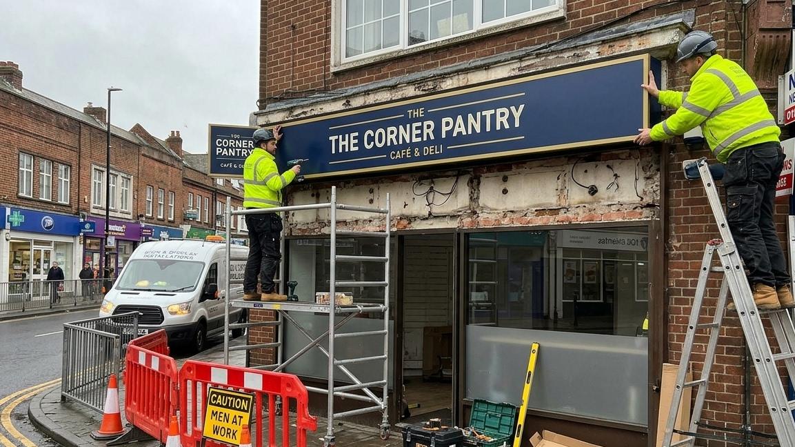 Professional installers fitting a fascia tray sign above a UK shopfront using secure mounting equipment.