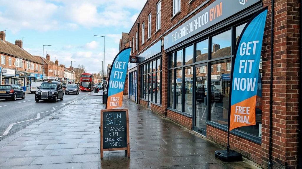 Two colourful feather flags and a wooden A-board pavement sign positioned outside a small gym in the UK, situated on a wide pavement with cars passing by on the road.