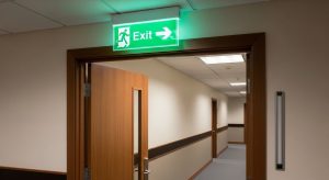 A well-lit and well-placed standard green fire exit sign with directional arrow mounted above a doorway in an office corridor.